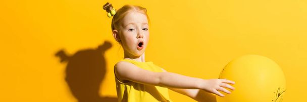 Girl with red hair on a yellow background. The girl is holding a yellow balloon on her outstretched arms. - Photo, Image