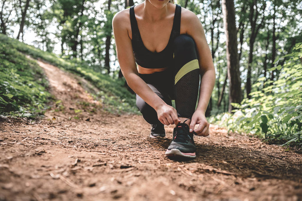 Cropped image of young athlete woman preparing to running outdoor on the forest path. Slim athletic girl ties up shoelaces on shoes before training in the park. - Fotoğraf, Görsel