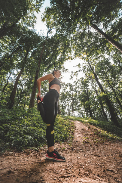 Low angle image of young woman athlete making warm-up before training in the woods. Side view of slim fit female preparing to run on forest path - Фото, зображення