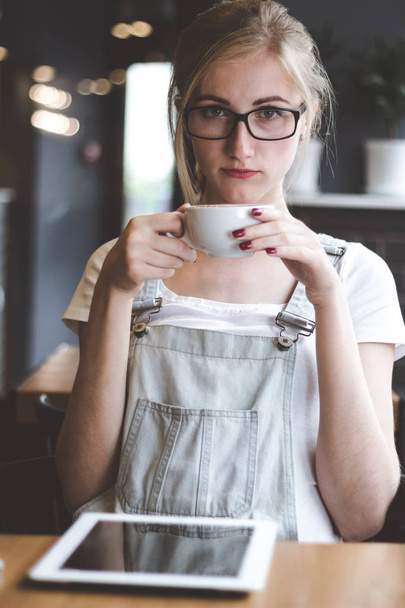 Enjoying her fresh coffee. Beautiful young woman drinking coffee and looking at camera while sitting in loft cafe. - Foto, Imagem