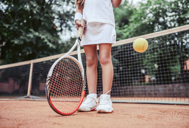 Summer activities for children in the tennis club. Cropped image of sports little girl on tennis court. Athletic child girl hits tennis ball with a racket. - Zdjęcie, obraz