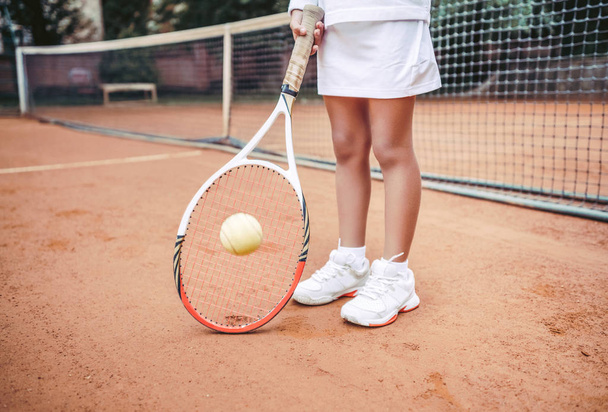 Child in sportwear playing tennis on outdoor court. Cropped image of child legs on tennis court. Close up view of tennis ball, racket and shoes. Active exercise for kids. - Photo, image
