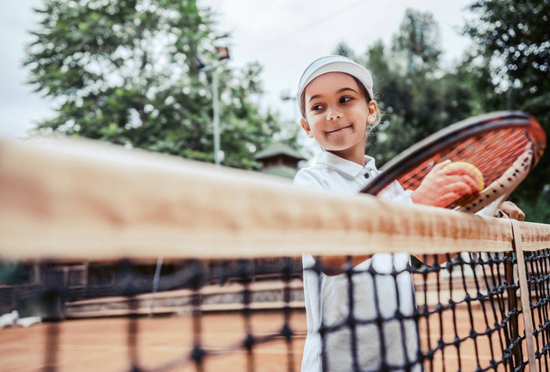 glückliches kleines Mädchen beim Tennisspielen. Kind in Sportkleidung hält Schläger und Ball an ein Tennisnetz gelehnt. - Foto, Bild