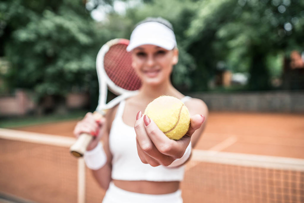 Beautiful young woman holding tennis racket and looking happy. Portrait of sportswoman with focus on tennis ball. - Foto, imagen