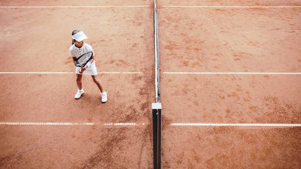 Child playing tennis on outdoor clay court. Top view of little tennis player on open tennis court. Full length shot of sporty child girl on tennis training in the club. Active exercise for kids. - Photo, image