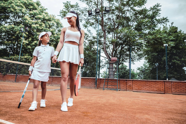 That was a great game! Full length image of sporty girl and woman holding hands after tennis match. Two happy tennis players on court. Family training. - Foto, afbeelding