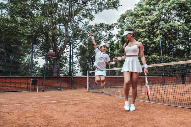 Little champion on tennis court! Full length image of jumping child tennis player with sporty trainer. Tennis coach holds hand of little girl celebrating victory. Active leisure outdoors. - Fotografie, Obrázek