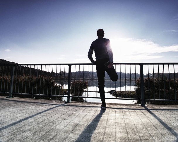 Man is stretching legs in break from run.  The Sun is outlining man body. Outdoor exercising on smooth concrete ground on lake bridge. - 写真・画像