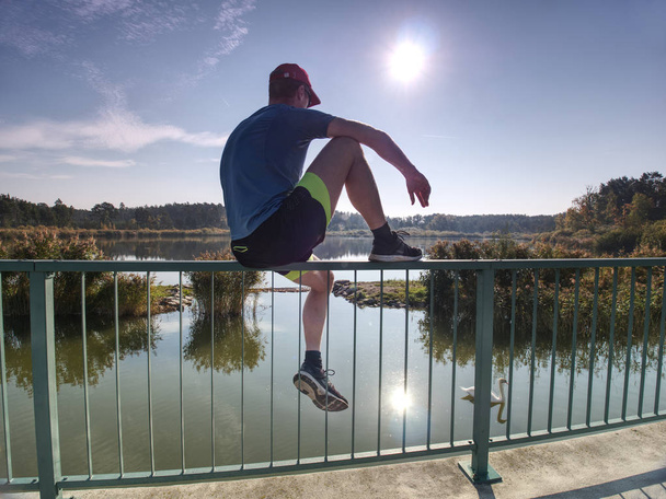 Ember a streching lábak a Futtatás szünetet. A Sun bemutatja az ember teste. Szabadtéri tó hídon sima beton alapon gyakorlása. - Fotó, kép