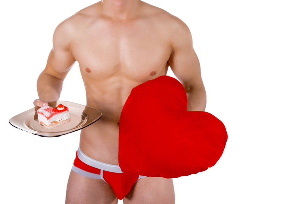 Torso of young handsome guy with heart pillow and heart-shaped cake on a plate. Sexy portrait of romantic man with valentine - 写真・画像