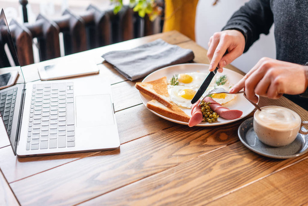 El hombre come. Cuchillo y tenedor en la mano. El concepto de comer en el trabajo. Laptop y desayuno en la mesa.. Desayuno de estilo americano con huevos fritos, salchichas, guisantes verdes y tostadas
. - Foto, Imagen