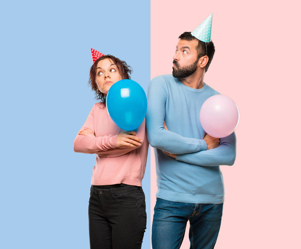 casal com balões e chapéus de aniversário fazendo gesto sem importância ao levantar os ombros sobre fundo rosa e azul
 - Foto, Imagem
