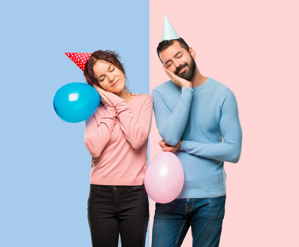 couple with balloons and birthday hats making sleep gesture. Adorable sweet expression on pink and blue background - Photo, Image