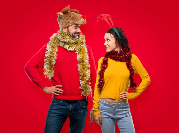 Couple dressed up for the christmas holidays posing with arms at hip and smiling on isolated red background - Фото, изображение