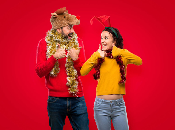 Couple dressed up for the christmas holidays giving a thumbs up gesture and smiling because has had success on isolated red background - Photo, Image
