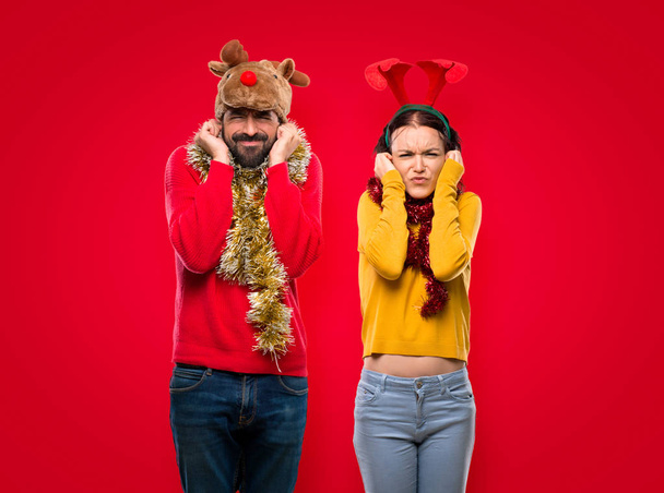 Couple dressed up for the christmas holidays covering ears with hands. Frustrated expression on isolated red background - Foto, Imagem