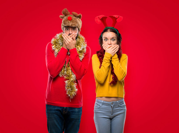 Couple habillé pour les vacances de Noël couvrant la bouche pour avoir dit quelque chose d'inapproprié. Ne peut pas parler sur fond rouge isolé
 - Photo, image