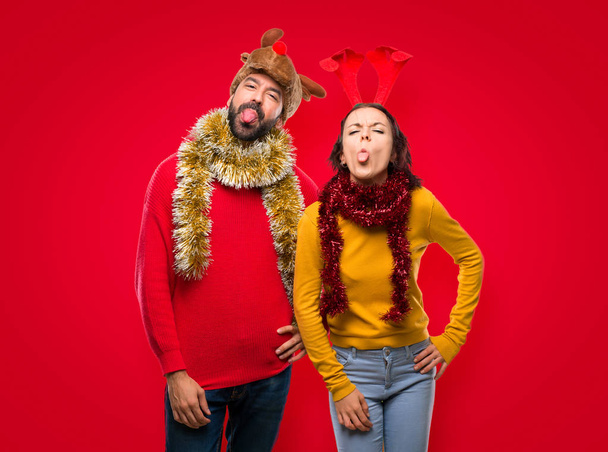 Pár oblečená pro vánoční svátky, zobrazující jazyk na kameru s legrační vypadají na izolované červeném pozadí - Fotografie, Obrázek