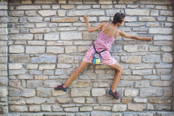 Eine Frau im Kleid klettert eine Ziegelmauer hinauf. klettert das Mädchen auf einen Steinzaun. Kletterer auf einer Stadtstraße. die Mauer des alten zerstörten Gebäudes. - Foto, Bild