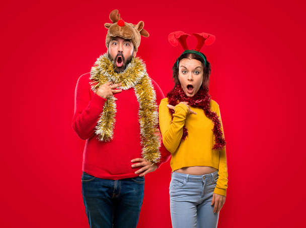 Пара, одягнена на різдвяні свята, здивована і шокована. Виразні емоції на обличчі на ізольованому червоному тлі
 - Фото, зображення