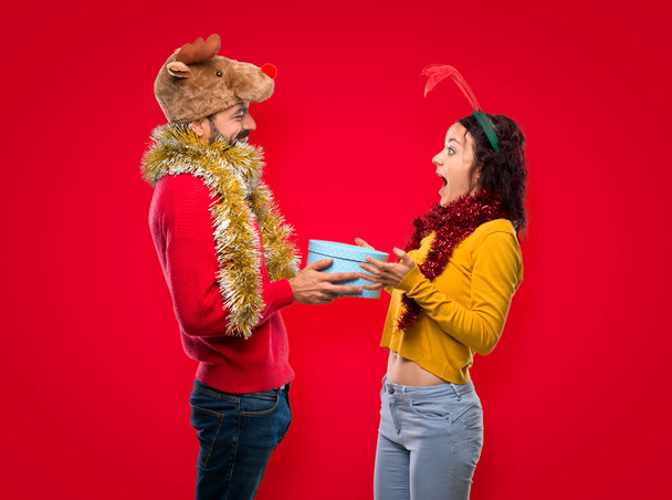 Couple dressed up for the christmas holidays holding a present on isolated red background - Photo, Image