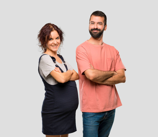 Para z kobieta w ciąży, utrzymując ręce skrzyżowane w pozycji bocznej, jednocześnie uśmiechając się na na białym tle szary - Zdjęcie, obraz