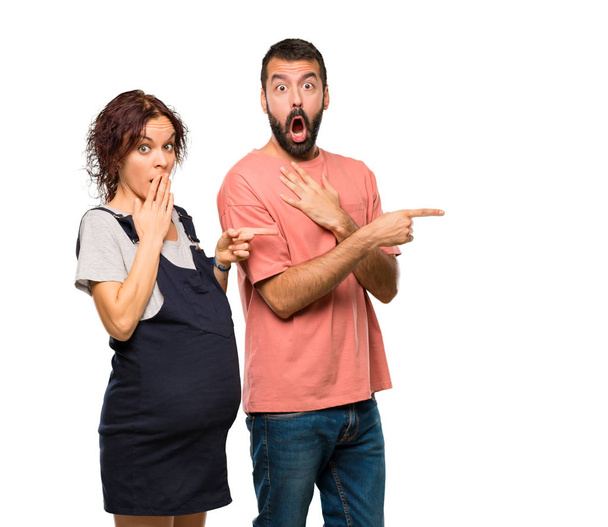 Couple with pregnant woman pointing finger to the side with a surprised face on isolated white background - Foto, imagen
