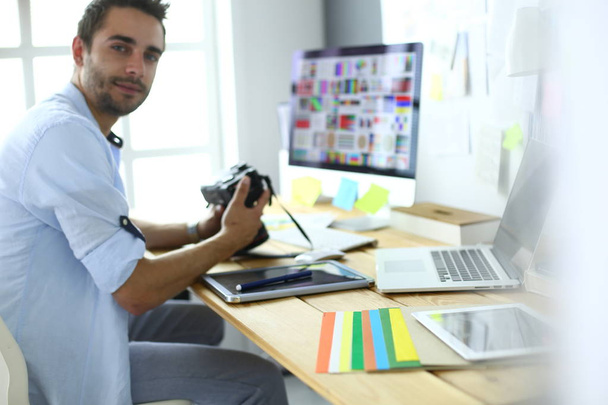 Retrato del joven diseñador sentado en el estudio gráfico frente a la computadora portátil y el ordenador mientras trabaja en línea. - Foto, Imagen