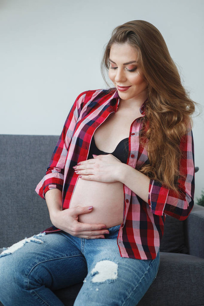 Mulher grávida bonita com cabelos longos em uma camisa xadrez está sentado em um sofá com uma barriga nua, acariciando suas mãos. Conceito de maternidade feliz, estilo de vida saudável
. - Foto, Imagem