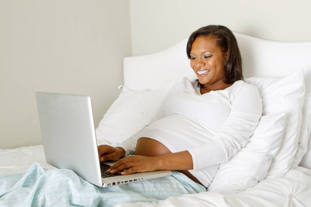 Young African American pregnant woman in bed on a computer. - Foto, Bild
