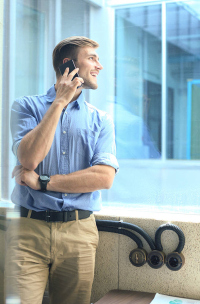 Uomo d'affari sorridente in piedi e utilizzando il telefono cellulare in ufficio
. - Foto, immagini