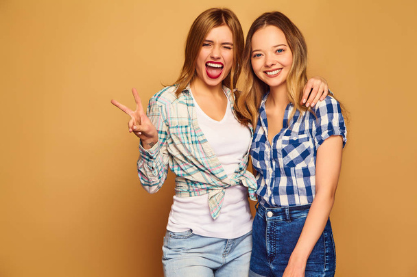 Two young beautiful blond smiling hipster girls posing in trendy summer checkered shirt clothes. Carefree women isolated on golden background. Positive models going crazy and hugging - Foto, immagini