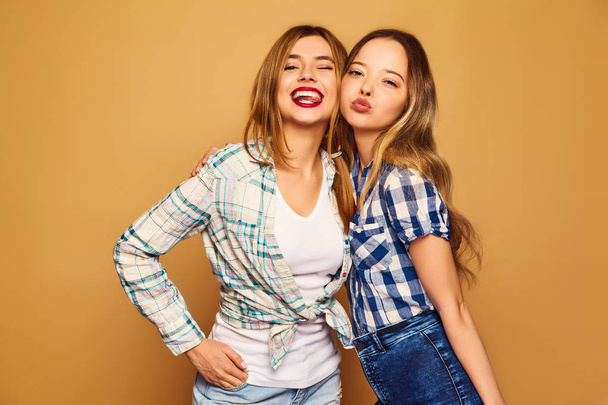 Two young beautiful blond smiling hipster girls posing in trendy summer checkered shirt clothes. Carefree women isolated on golden background. Positive models going crazy and hugging - Фото, зображення