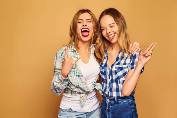 Two young beautiful blond smiling hipster girls posing in trendy summer checkered shirt clothes. Carefree women isolated on golden background. Positive models going crazy and hugging - Fotó, kép