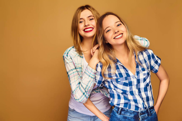 Дві молоді красиві біляві усміхнені дівчата-хіпстери позують у модному літньому картатому одязі. Безтурботні жінки ізольовані на золотому тлі. Позитивні моделі божевільні і обіймаються
 - Фото, зображення