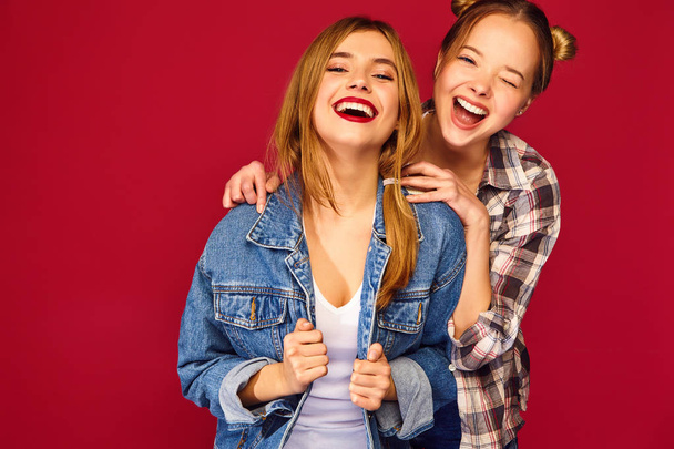 Two young beautiful blond smiling hipster girls posing in trendy summer checkered shirt clothes. Carefree women isolated on red background. Positive models going crazy and hugging - Foto, Imagem