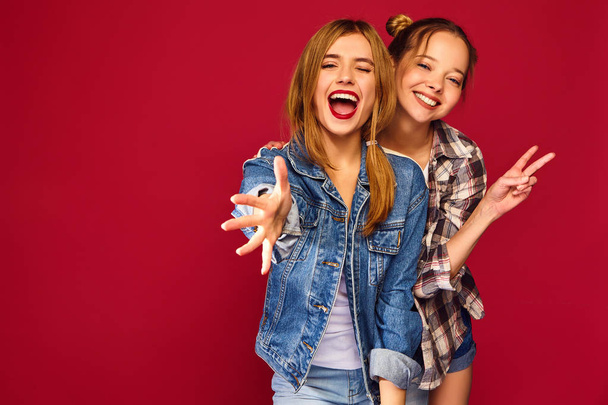 Two young beautiful blond smiling hipster girls posing in trendy summer checkered shirt clothes. Carefree women isolated on red background. Positive models going crazy and hugging - Foto, immagini