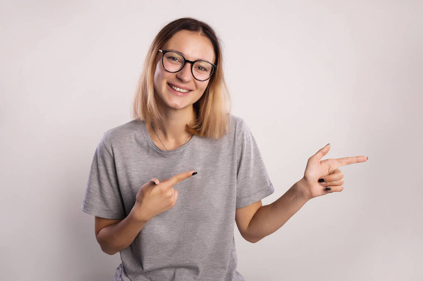 Genç kadın gülümsüyor ve işaret veya gösteri yön. Kız büyük gözlük ve gri arka plan kopya alanı üzerinde iki eliyle kamera ve puan parmak bakmak gri gömlek - Fotoğraf, Görsel