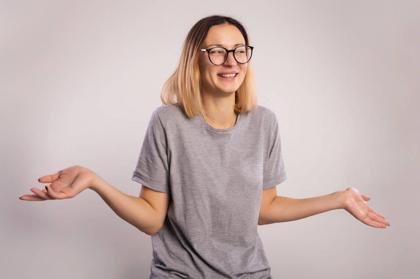 I don't know concept. Portrait of a young woman who do not understand what is happening. The girl in big eyeglasses and gray shirt raises hands and shrugging his shoulders on a gray background copy space - Photo, Image