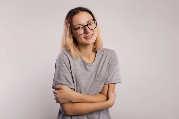 Portrait of a casual woman in big glasses and gray shirt looking at camera and hugging herself isolated on a gray background - Фото, зображення
