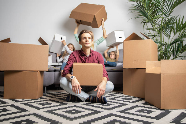 Photo of serious man sitting on floor and women with boy and girl sitting on gray sofa among cardboard boxes - Фото, изображение
