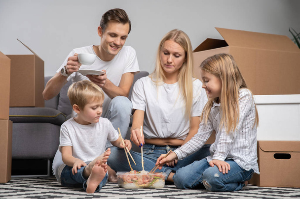 Εικόνα των γονέων με τα παιδιά να τρώνε ρύζι με γαρίδες που κάθεται στο πάτωμα ανάμεσα σε κουτιά από χαρτόνι - Φωτογραφία, εικόνα