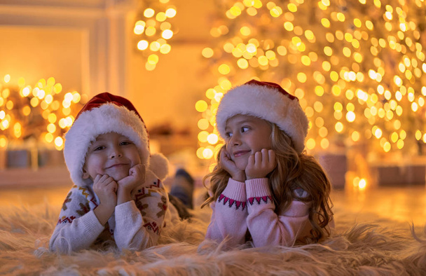 Feliz Navidad y Feliz Año Nuevo! Niños esperando vacaciones de invierno en casa. Hermano pequeño y hermana en los sombreros de Santa tendidos en el suelo cerca del árbol de Año Nuevo decorado por la noche
. - Foto, imagen