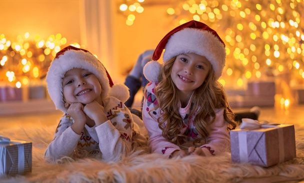 Merry Christmas and Happy New Year! Children waiting for winter holiday at home. Little brother and sister in Santa hats lying on floor near decorated New Year tree in the evening. - Foto, imagen