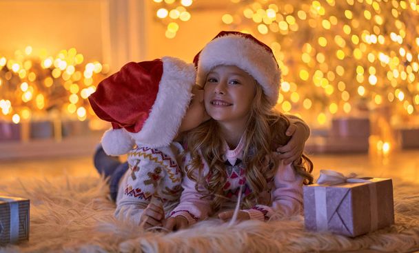 Merry Christmas and Happy New Year! Children waiting for winter holiday at home. Little brother and sister in Santa hats lying on floor near decorated New Year tree in the evening. - Fotografie, Obrázek