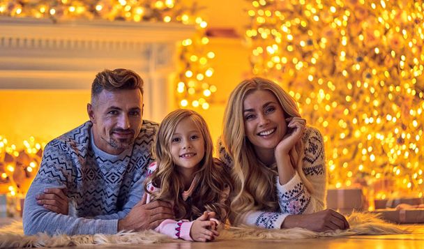 Merry Christmas and Happy New Year! Happy family celebrating winter holiday at home. Mother, father and daughter are waiting for Christmas while lying near decorated New Year tree. - Foto, imagen