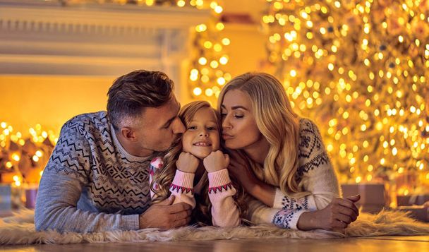 Merry Christmas and Happy New Year! Happy family celebrating winter holiday at home. Mother, father and daughter are waiting for Christmas while lying near decorated New Year tree. - Foto, Imagen