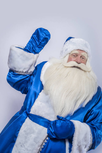 Santa Claus y el padre Frost con un abrigo azul están de pie sobre un fondo blanco aislado. Un anciano, un patriarca y un mago están mirando a alguien o algo.
. - Foto, Imagen