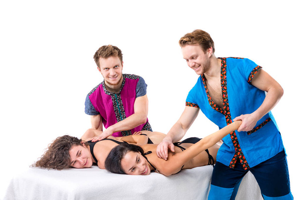 Massage à thème et soins du corps. Deux jeunes frères jumeaux mâles avec une barbe et un uniforme font un massage en tandem dans les mains de deux femmes caucasiennes sexy allongées sur une table sur un fond blanc isolé
. - Photo, image