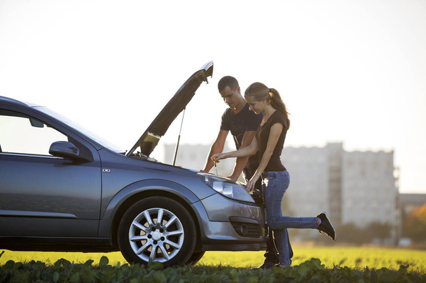 Young couple, handsome man and attractive woman at car with popped hood checking oil level in engine using dipstick on clear sky background. Transportation, vehicles problems and breakdowns concept. - Photo, Image